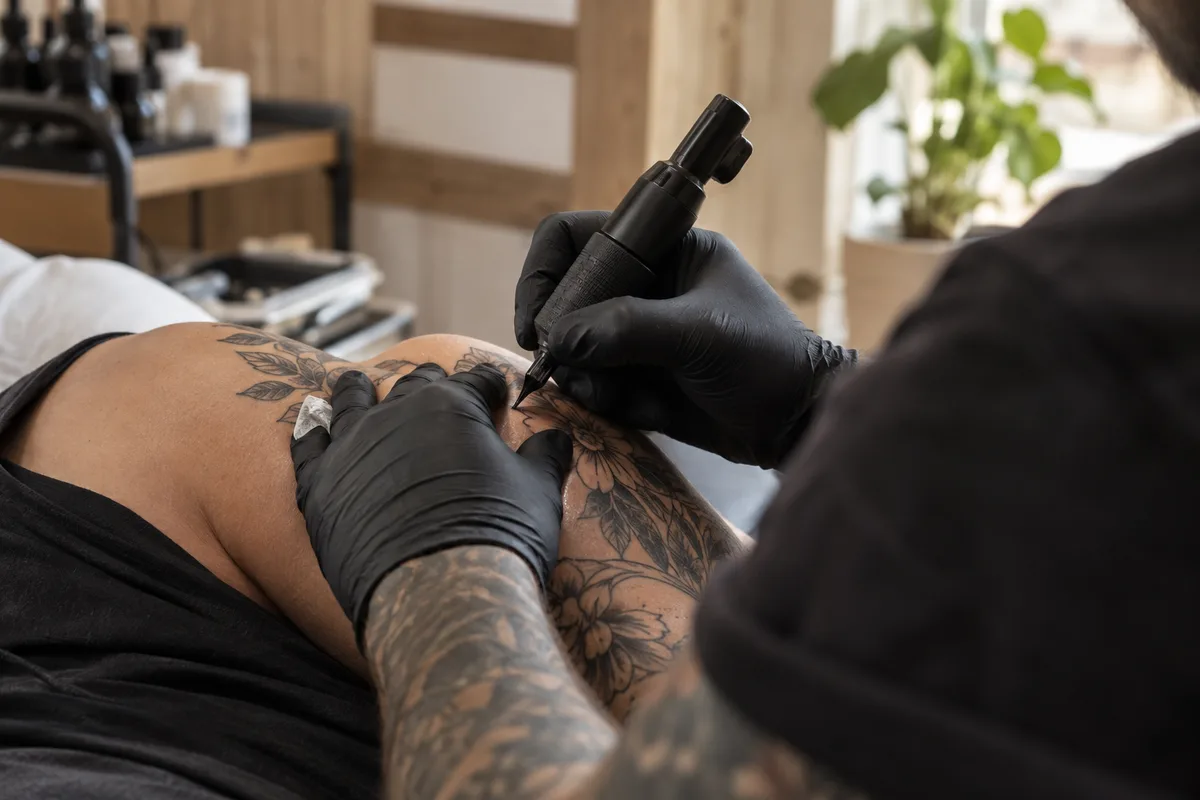 Tatoueur vu de dos penché sur une épaule, mains gantées et machine noire unie
